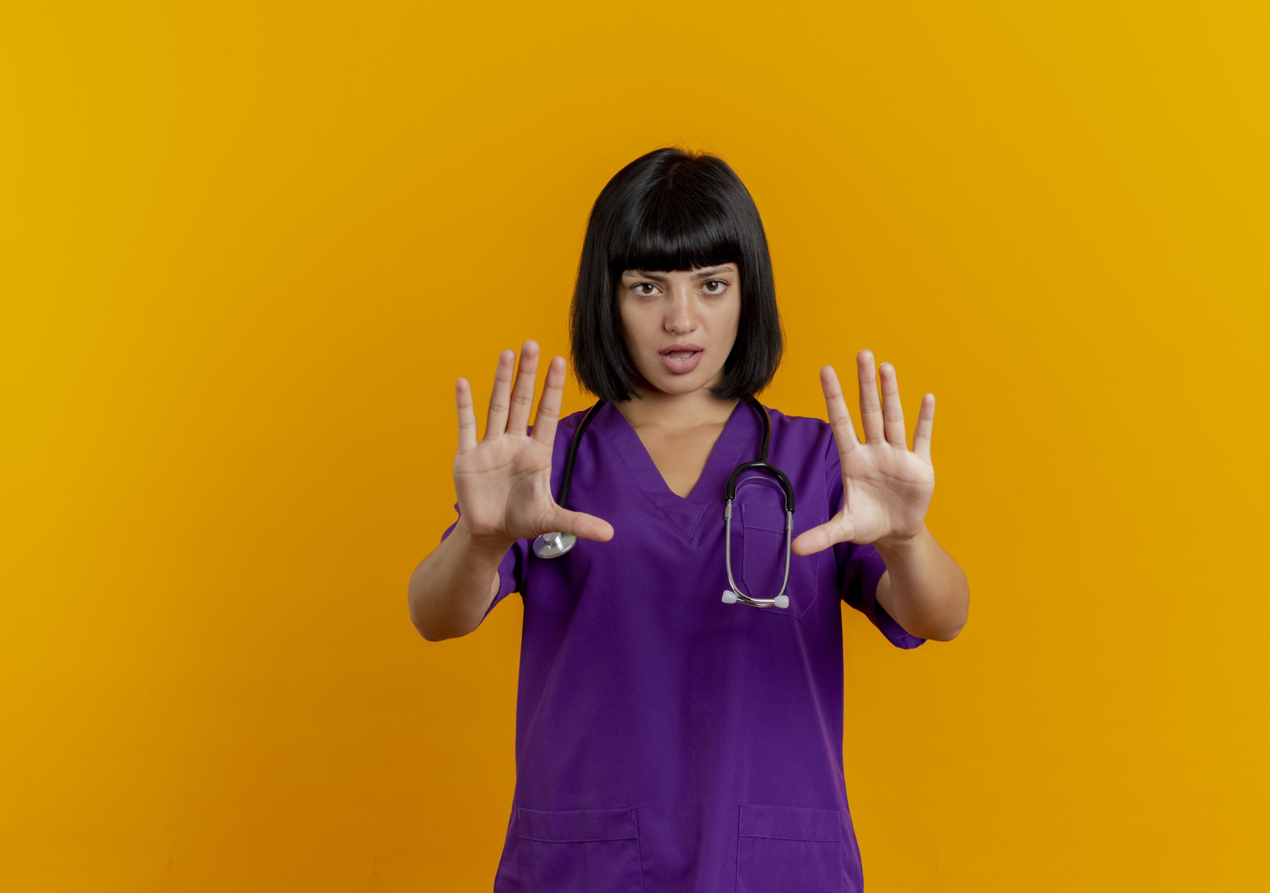 confident young brunette female doctor in uniform with stethoscope gestures stop hand sign with two hands isolated on orange background with copy space